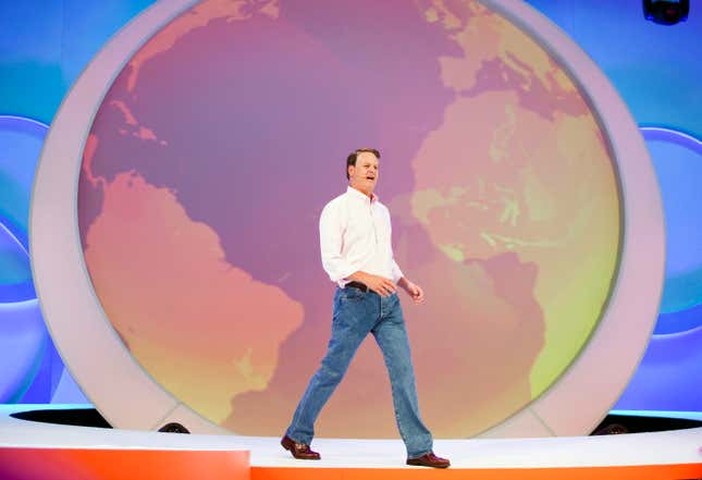 Nike CEO John Donahoe (in a white button down and jeans) stands and speaks on stage in front of a globe