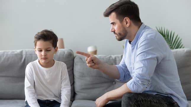 A man sits on a couch and sternly points his finger at his son who looks away. 