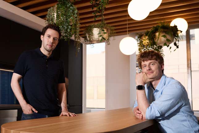 one man wearing a black t shirt is standing with his hand placed on a table, across from him another man wearing a blue button down is sitting with his chin rested on his arm which is also on the table