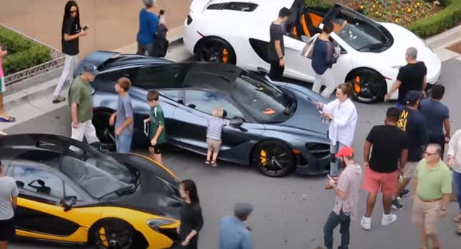 A blond headed kid leans on a McLaren and breathes on it, probably getting little blond kid germs all over it. Definitely bad for the paint.