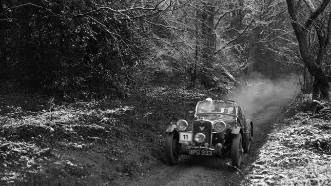 A black and white photo of a vintage car driving on a snow-covered trail. 