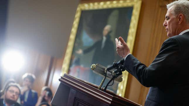 Kevin McCarthy at a podium in a press conference room facing attendees and a portrait of George Washington hanging on the wall