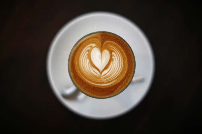 A latte in a white cup and saucer with a black background and heart in the foam on top.