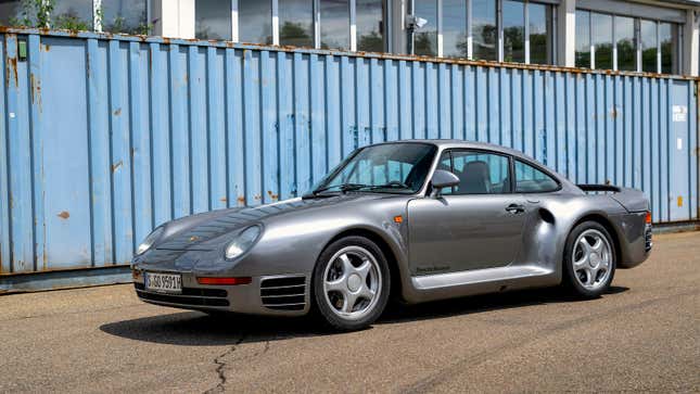 A dark silver Porsche 959 is parked in front of a shipping container.