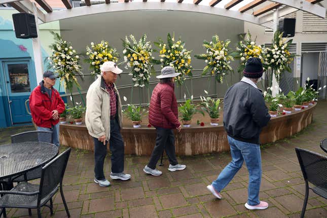 On the one-year anniversary of a mass shooting, a group of people walk past a display of flowers memorializing the victims, on a stage at Mac Dutra Plaza in Half Moon Bay, Calif., Tuesday, Jan. 23, 2024. (Scott Strazzante/San Francisco Chronicle via AP)