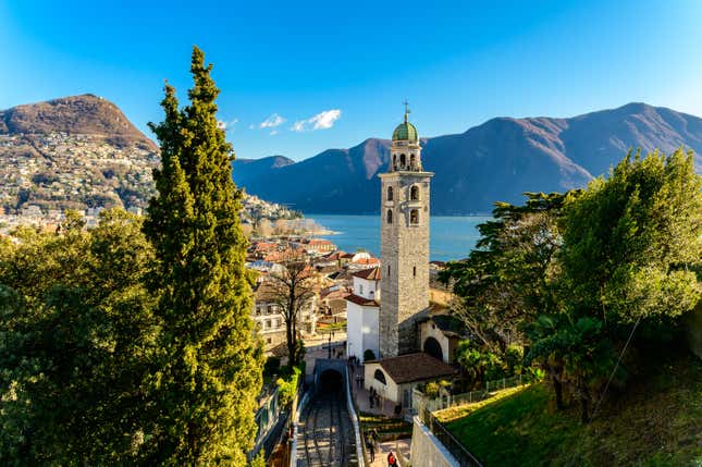 aerial view of old buildings and a church clocktower on a sunny day with a body of water and mountains in the background