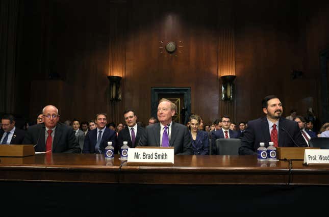 Three men sitting at a table, wearing suits, with name plates in front of them, the middle one says Mr. Brad Smith.