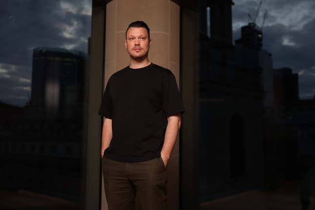 Victor Riparbelli wearing a black t shirt and olive green pants, his hands are tucked in his pants and he is looking up