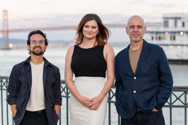 three people stand side by side on a waterfront with a bridge faded in the background behind them