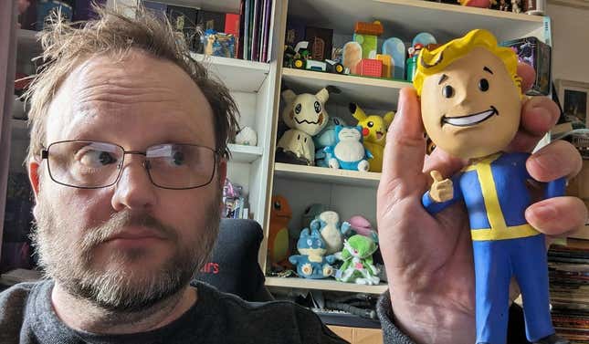A man who needs a haircut holds a Fallout Vault Boy bobblehead by its broken head, shortly after gluing its thumb back on.