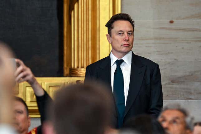 Elon Musk standing and looking at something or someone, people around him appear to be sitting and are blurry