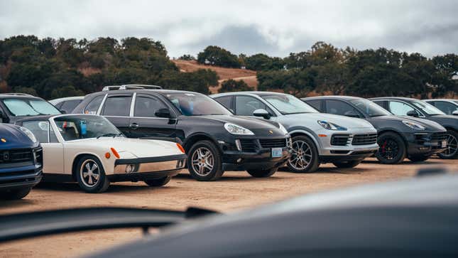 A white Porsche 914 is parked on dirt among a bunch of large Cayenne SUVs