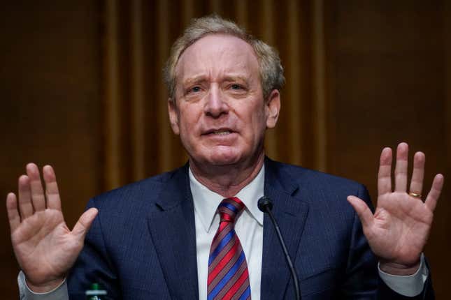 Brad Smith wearing a white button down and striped tie with navy suit jacket raising both hands as he speaks at a microphone