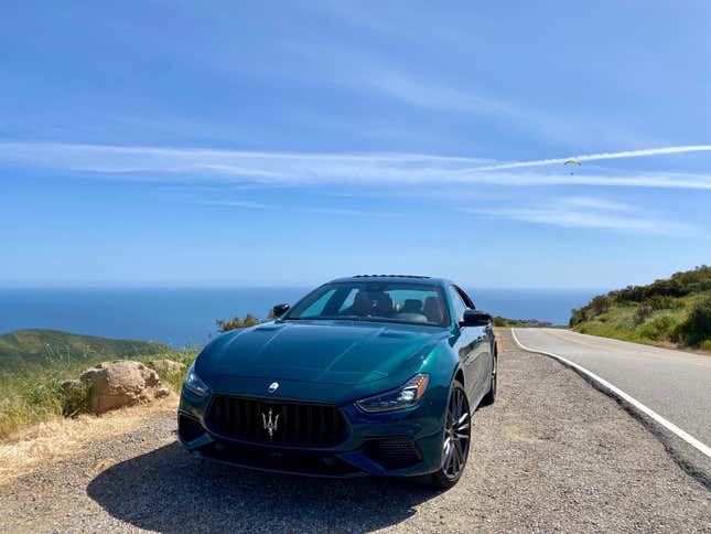 A front shot of the car parked on a mountainside with the ocean and a hang glider in the distance