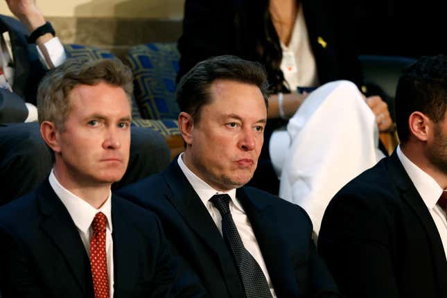 Elon Musk sits in a black suit surrounded by other people also wearing suits