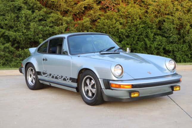 A silver 1974 Porsche Carrera 2.7 is parked in front of some bushes