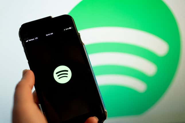 A hand holds a smart phone up, and the screen displays the Spotify logo. In the background, another Spotify logo.