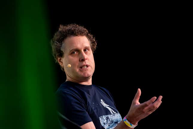 Peter Beck wearing a black t-shirt and speaking with a microphone he's wearing on his head with his hand up