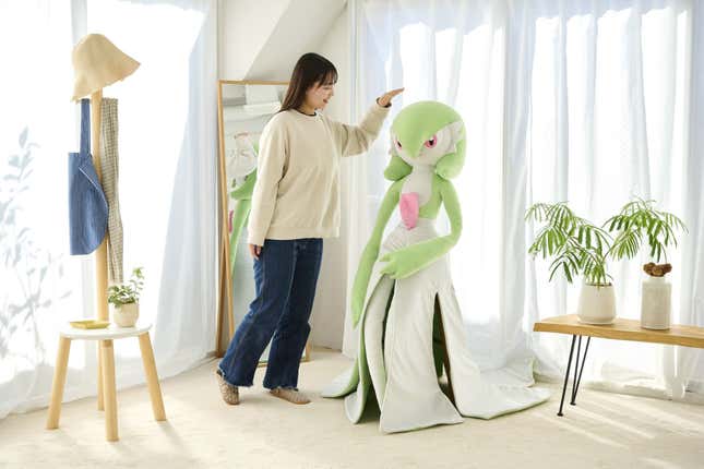 A person is shown standing next to the Gardevoir plush and patting it on the head.