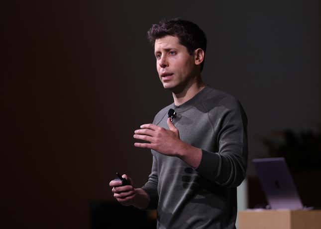 Sam Altman wearing a grey sweater and speaking with his hand up on stage