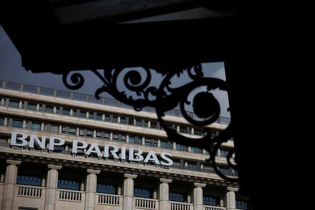 A view of a BNP Paribas bank. In the foreground is a building with curly ironwork under its eaves, set in silhouette.