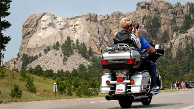 A photo of two people riding a motorbike near mount Rushmore. 