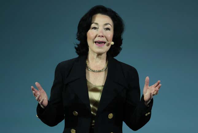 Safra Catz wearing a gold shirt and black blazer over it speaking with her hands up in front of a bluish grey backdrop