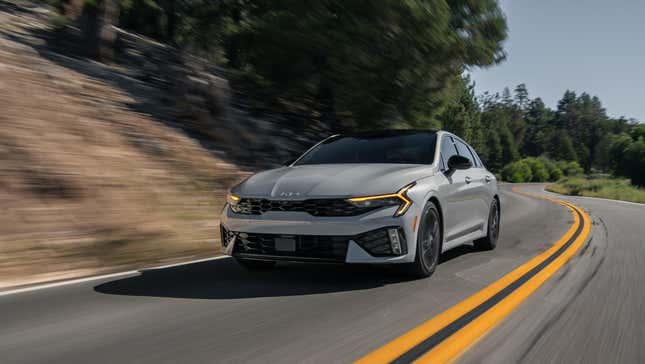 a gray Kia K5 driving on a two-lane road in front of trees