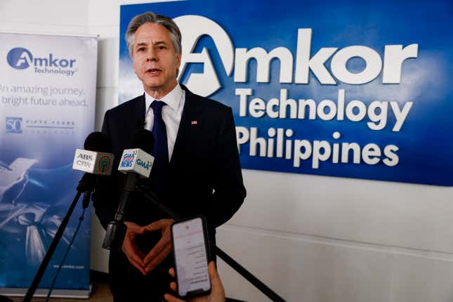 antony blinken speaking in front of a sign with Amkor Technology Philippines logo, two microphones and a hand holding a phone are in front of him