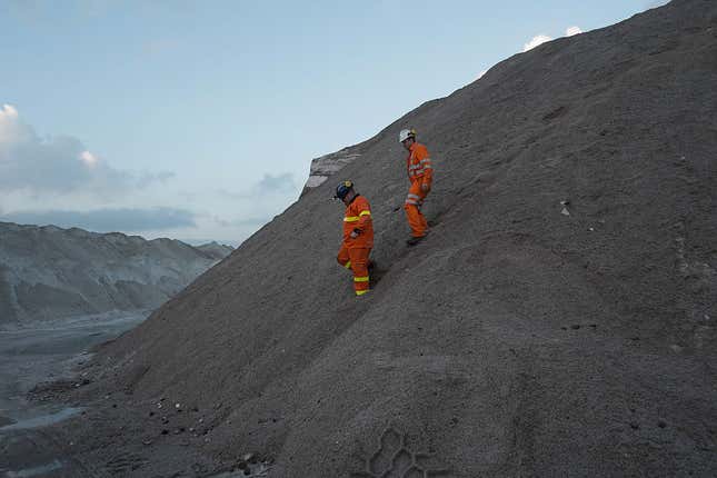 Two people in orange protective suits walk down a huge gray slope of potash.