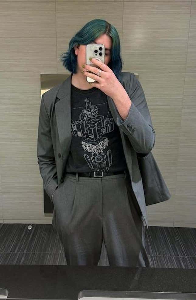 Bathroom selfie of a woman with blue hair wearing a gray suit and a transitor t-shirt