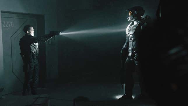A soldier points their pistol as Master Chief in a dark room. 
