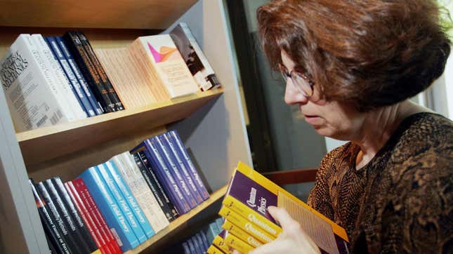 A woman holds a stack of yellow textbooks next to a bookshelf with a space laid out next to quantum mechanics.