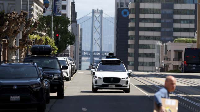 A white self-driving car drives down a San Francisco Street with the Bay Bridge in the background. 