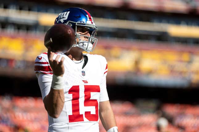 Giants' QB, wearing a blue helmet and white jersey with red numbers, spins a football in his finger in a large football stadium. The seats are vaguely visible behind him, in alternating sections of red and yellow.