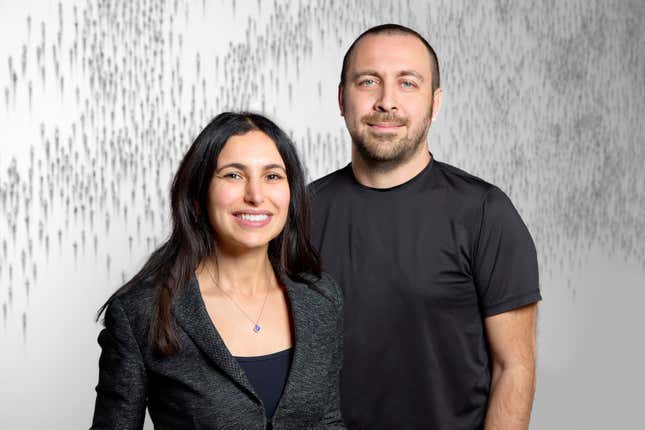 a woman is wearing a black shirt and grey blazer over it sitting and smiling while a man stands beside her in a black tshirt also smiling