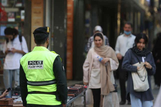 Foreground: An Iranian police officer on patrol. Background: Two Iranian women, one wearing a headscarf, one not.