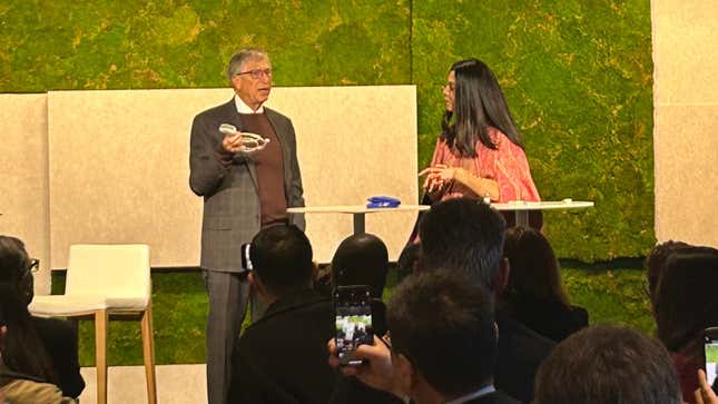 Bill Gates holds a medical device on stage as an interviewer looks on