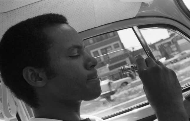 A black and white photo of a man lighting a cigarette inside what looks like a classic VW Beetle using the cigarette lighter