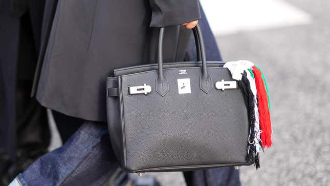 A photo of a black Hermes bag held by a person in a black trenchcoat and raw denim jeans.