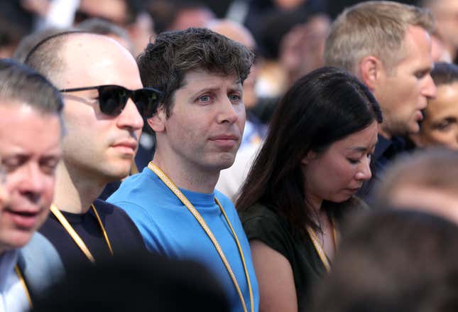 Sam Altman wearing a blue shirt and orange lanyard, looking onward in a crowd of people