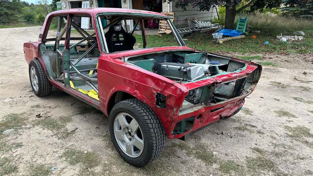 Un sedán Lada rojo está estacionado, sin puertas, en un patio con un asiento de carreras y una jaula antivuelco instalada