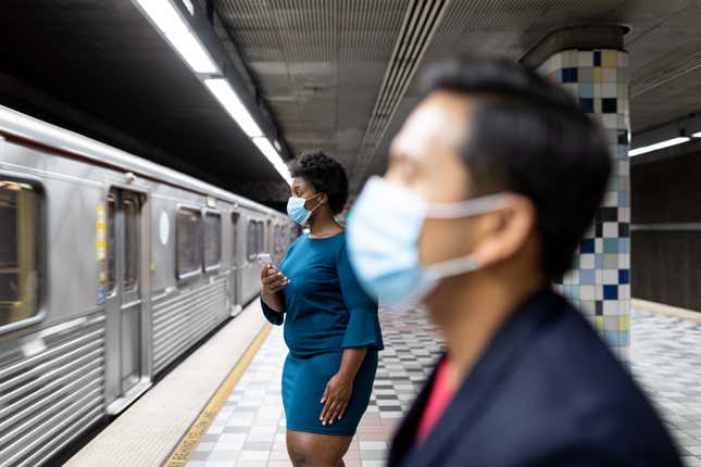 Two people wearing surgical masks wait at a subway station.