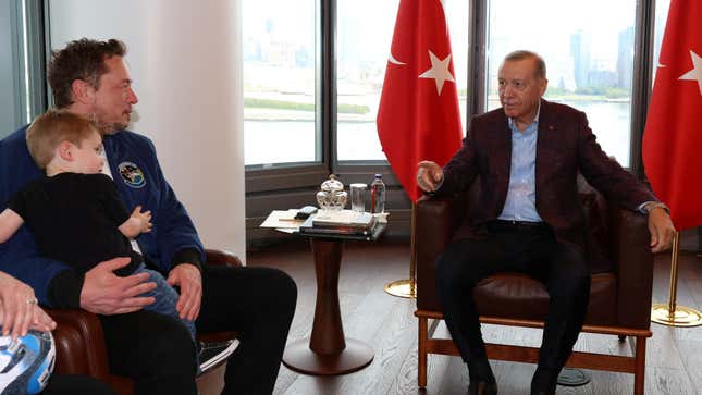 Elon Musk, holding a child in his lap, and Turkish president Recep Tayyip Erdoğan sit chairs next to windows and two Turkish flags.
