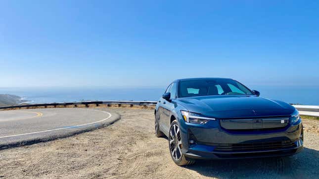 A navy blue Polestar 2 from the front 3/4 angle parked on dirt on a cliff in Malibu with the coastline in the distance