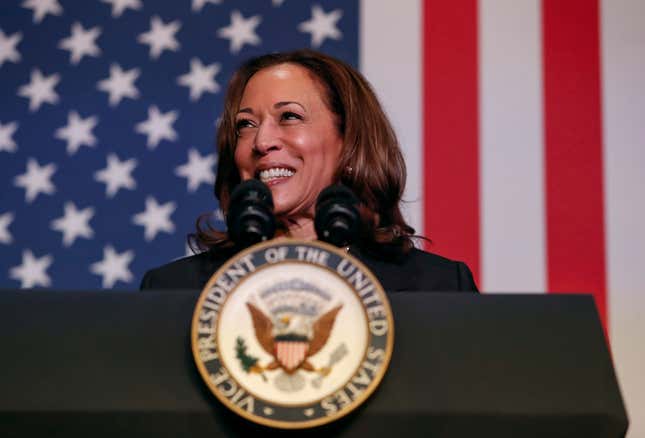 Kamala Harris lächelt hinter einem Podium mit dem Siegel des Vizepräsidenten der Vereinigten Staaten, hinter ihr weht eine amerikanische Flagge