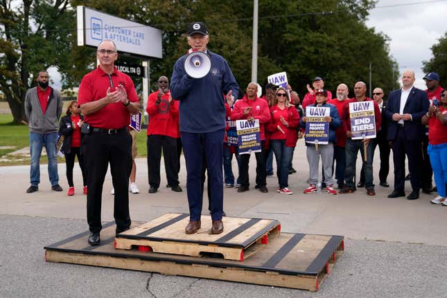 File - United Auto Workers President Shawn Fain, left, listens as President Joe Biden speaks to striking UAW members outside a General Motors facility on Sept. 26, 2023, in Van Buren Township, Mich. Throughout its 5-week-old strikes against Detroit’s automakers, the United Auto Workers union has cast an emphatically combative stance, reflecting the style of Fain, its pugnacious leader. (AP Photo/Evan Vucci, File)