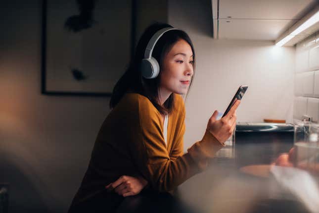 Asian woman leaning against kitchen counter looking at phone, smiling, with overhead earphones on