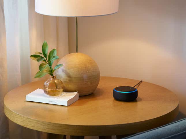 Echo Dot sentada en una mesa de madera marrón claro junto a un cordero, un libro y una planta artificial