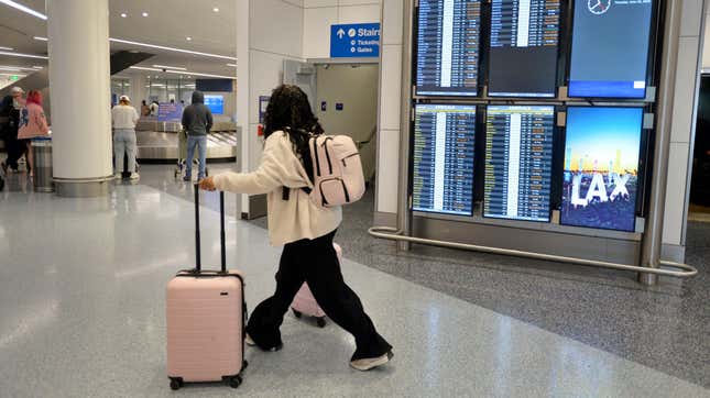 Un pasajero camina por un aeropuerto junto al panel de llegadas y salidas empujando maletas de color rosa claro